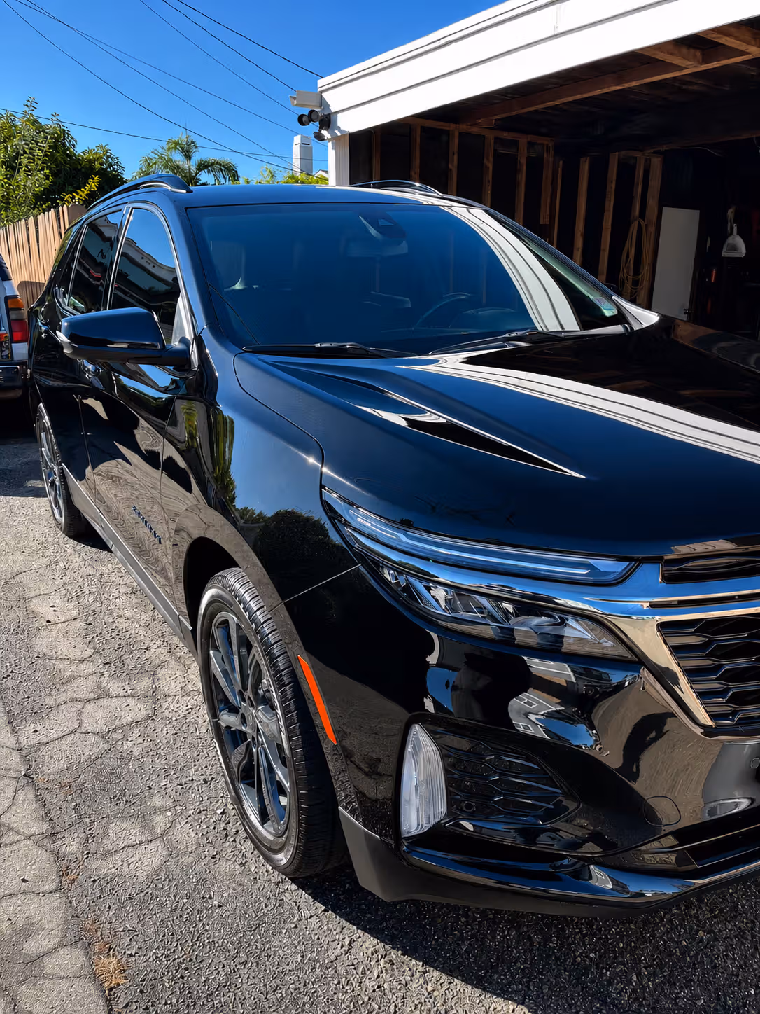 Black modern SUV parked on a sunlit driveway beside a wooden fence and carport.
